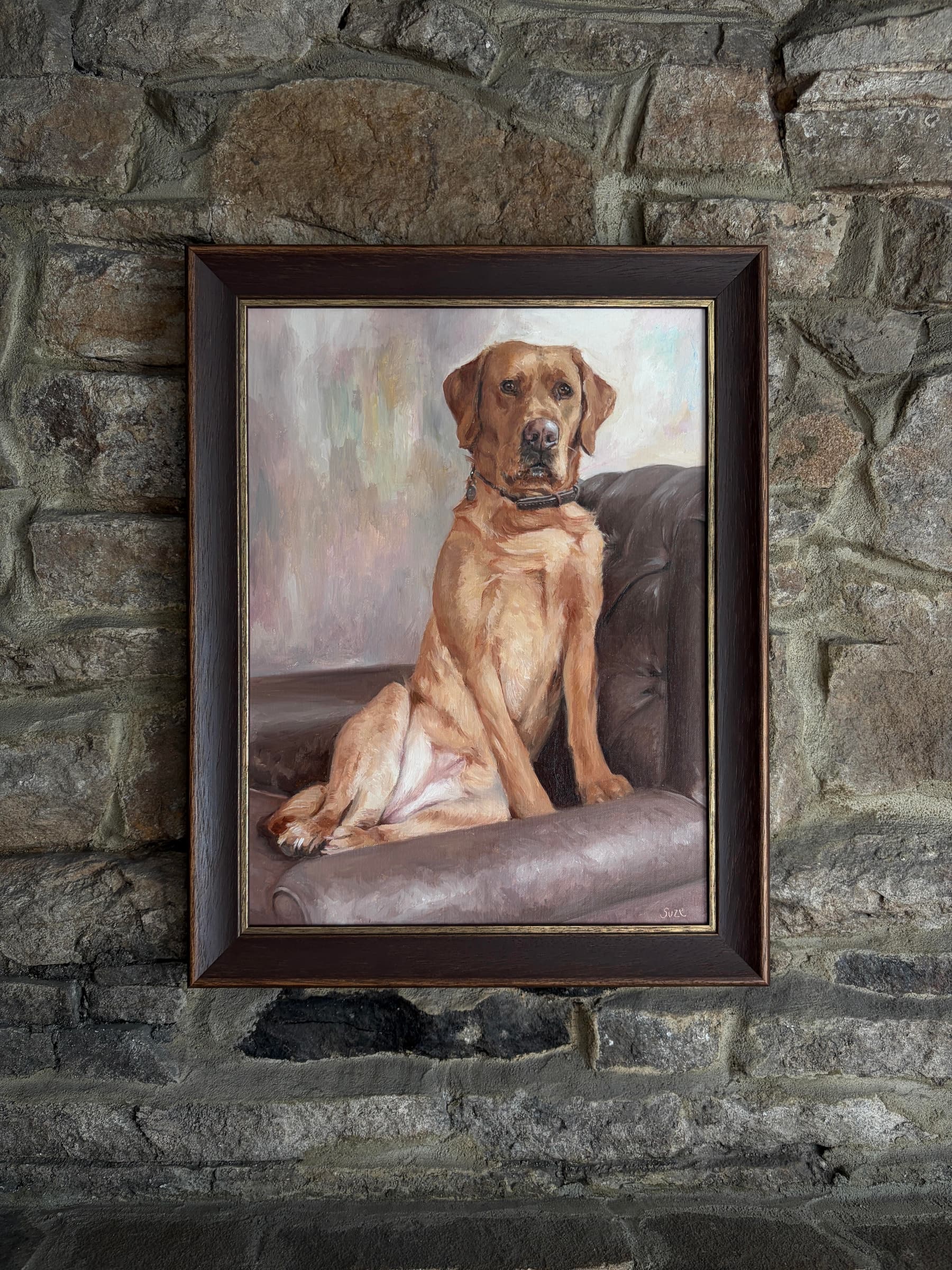 A framed painting of a golden-coated dog seated on a brown armchair, displayed against a stone wall